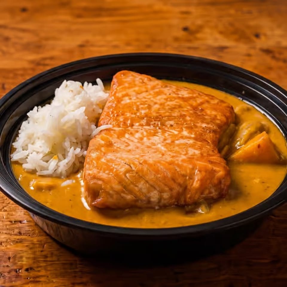 Salmon fillet with white rice and yellow curry in a black bowl on a wooden table.